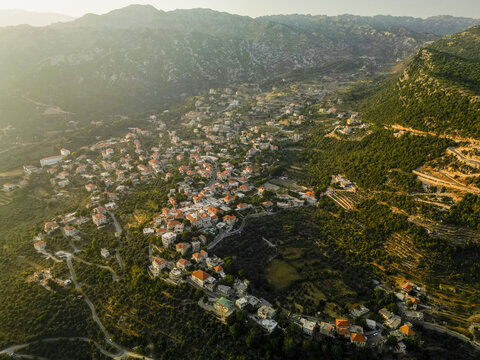 Aerial view of a cluster of homes with red roofs nestled amidst the undulating hills and verdant forests, bathed in the golden light of the sun, Douma, North Governorate, Lebanon.