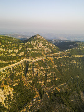 Aerial view of a winding road slicing through the rugged, sun-drenched mountains, where the vibrant green of dense forests meets the stark, rocky peaks, Douma, North Governorate, Lebanon.