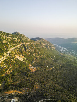 Aerial view of dramatic mountains rise with winding roads carved into the steep slopes, shrouded in lush greenery under a soft sky, Douma, North Governorate, Lebanon.