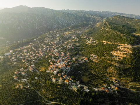 Aerial view of a village nestled among the rugged, sun-drenched mountains, where terracotta roofs meet verdant hillsides under a hazy sky, Douma, North Governorate, Lebanon.