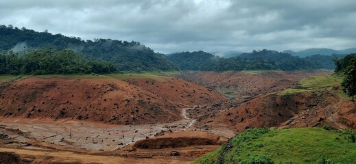 Dry, exposed reservoir bed at Anathode flanking dam revealing cracked soil, tree stumps, muddy channels, with lush green forested hills, mist-covered mountains in background under sky in kerala, india