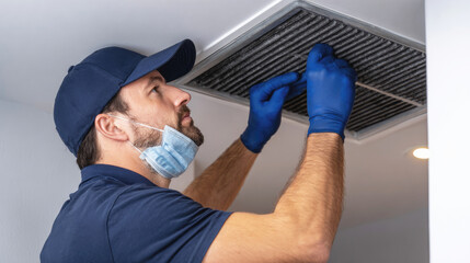HVAC technician wearing gloves and mask cleaning dirty air duct filter indoors, maintaining ventilation system and ensuring clean airflow in residential or commercial setting.