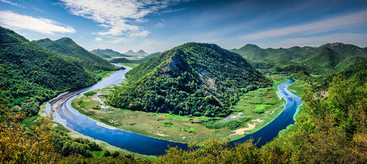 Panoramic view of a meandering river bend through lush green mountains under clear blue sky; serene summer landscape, crystal water, vibrant nature, European travel destination.