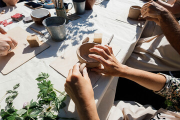 Hands carefully shape clay into small bowls at a pottery making workshop held outdoors. Participants are focused and enjoying the creative process surrounded by greenery