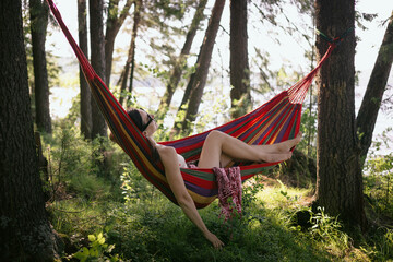 person enjoys leisure time in a striped hammock nestled between trees. bright sunlight filters through the leaves, casting playful shadows on the ground