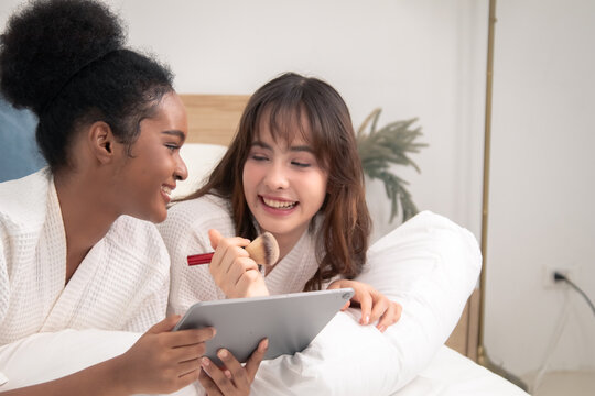 Two teenage friends lying on bed together, smiling while holding tablet and makeup brush, relaxed atmosphere showing natural healthy skin and cheerful bonding during leisure moment, different skin - Powered by Adobe