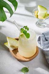 Refreshing lemonade or mojito in a glass with melon, straw and mint on a wooden plate on a light background