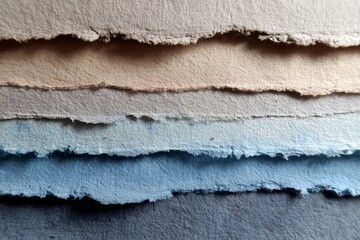 A close-up view of layered handmade papers, showcasing a spectrum of muted colors from beige to deep blues, with visible textures and frayed edges.