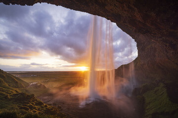View of golden light cascading down Seljalandsfoss waterfall framed by a dark cave opening onto a vibrant landscape under a dramatic sky, Iceland, RangÃ¡rÃ¾ing eystra, Iceland.