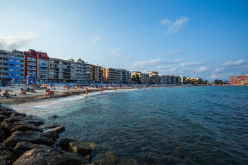 Fototapeta premium A panoramic view of Torrevieja’s coastline, showing a curved sandy beach lined with apartment buildings and the calm Mediterranean Sea. The scene captures a lively seaside atmosphere with people relax