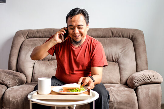 Man Sitting on Sofa Having a Phone Call While Holding Plate with Sandwich for Breakfast