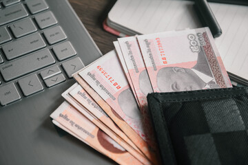 Cambodian Riel banknotes in a wallet on a laptop, with an open notebook and pen on a wooden desk. Represents online finance, digital transactions, budgeting, and planning.