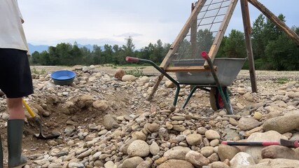 Outdoor adventures on river. Gold panning. A gold prospector is dry sifting sand and gravel from a river using a shovel, a homemade sieve, and a wheelbarrow. The sand will be washed into a sluice box 