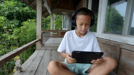 A boy in headphones uses a tablet for online learning while sitting on a rustic porch, illustrating a modern digital nomad family lifestyle and the concept of remote education - Powered by Adobe