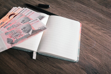 Cambodian Riel banknotes arranged on a wooden desk, next to an open notebook and pen. This image...