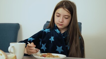 Bowl of buckwheat porridge being eaten by girl in kitchen with calm face. Child indoors holding fork while tasting simple homemade meal. Young person at table enjoying traditional buckwheat dish with