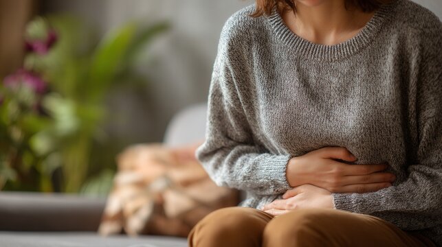A woman in a gray sweater holds her abdomen, suggesting discomfort, with a blurred indoor background of plants and a couch.