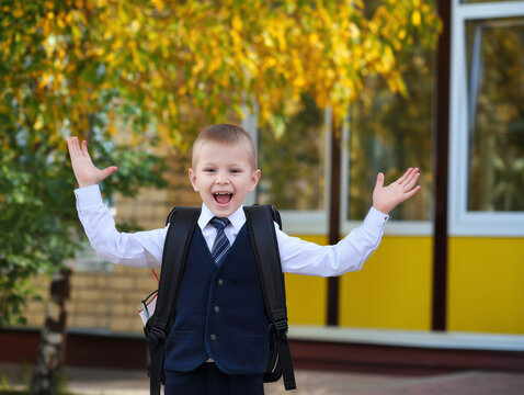A happy and excited young schoolboy in a blue uniform and with a backpack raises his hands in a gesture of joy and freedom, celebrating the first day of school on a sunny autumn day