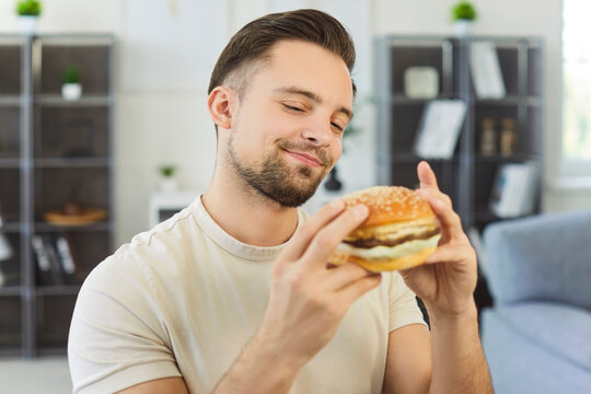 Young 25 Caucasian man holding juicy hamburger with hands and smiles with satisfaction, sitting comfortably alone at home. Fast food enjoyment, unhealthy eating and bad life habits concept