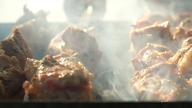 Extreme close-up juicy meat pieces on skewers roasting over hot coals. Grill master rotates kebab skewers for even cooking. Shiny spiced meat glistens in sunlight, releasing smoke and aroma.