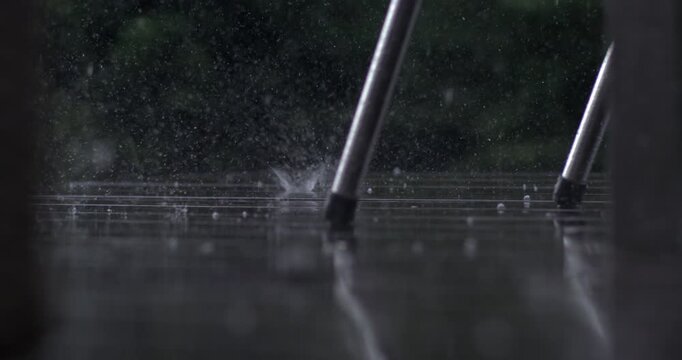 Low angle view of rain splashing around chair legs on wet wooden deck, captured in super slow motion with high speed camera, moody and reflective scene
