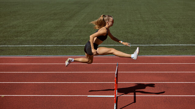 Female hurdler mid-air jumping hurdle on track. Concept of overcoming barriers, strength in motion, determination, resilience, and pushing limits in athletics. - Powered by Adobe