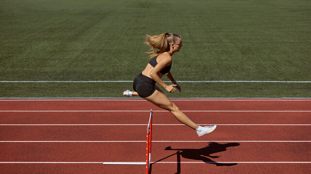 Female athlete mid-air jump over hurdle on stadium track. Concept of determination, overcoming obstacles, sport psychology, resilience, and powerful action moment.
