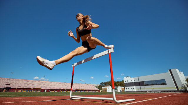 Female hurdler mid-air over hurdle with dynamic stride on stadium track. Concept of athletics, power, determination, and overcoming obstacles in sport and life. - Powered by Adobe