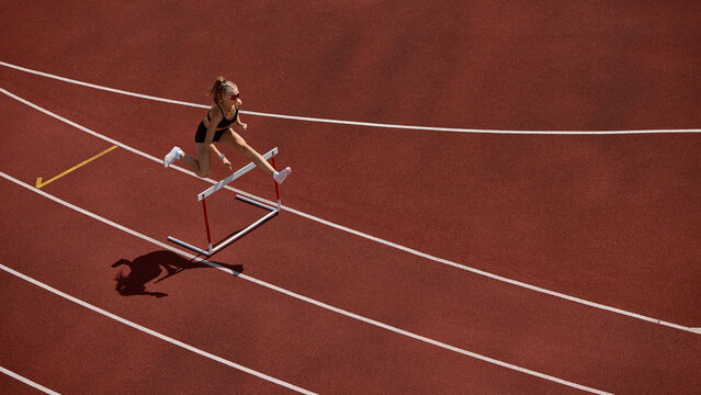 Female track athlete sprinting and clearing hurdle in powerful action motion. Concept of athletics, leadership in action, mental barriers, coaching for growth, and financial risk strategy.