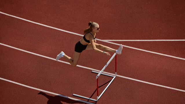 Female athlete clearing hurdle mid air on stadium track with determination. Concept of business resilience, leadership under pressure, coaching for success, and female empowerment.