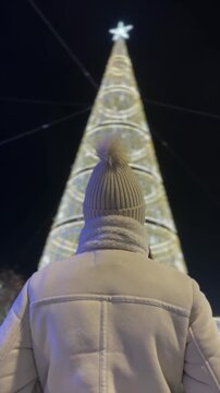 Woman in winter coat and wool hat looking at illuminated Christmas tree at night
