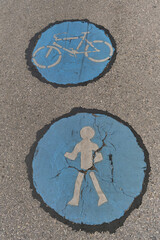 Detailed close-up of asphalt pavement featuring two round blue road markings with white icons — bicycle on top and pedestrian below — showing cracked and faded paint.