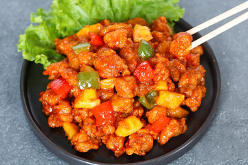 Sweet and sour chicken served on a dark plate, with a pair of chopsticks picking up a piece. Decorated with bell peppers and pineapple chunks. A classic Chinese comfort food.