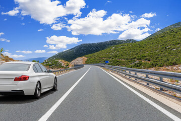 A white car drives along a scenic mountain road under a bright blue sky with fluffy clouds.