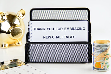 A desk displays a message of gratitude for embracing new challenges, alongside a calculator, a piggy bank, and rolled cash, symbolizing financial planning and success