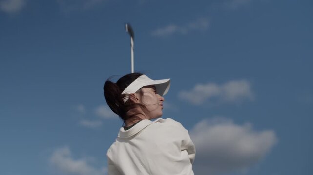 Female golfer with visor hat swings golf club against blue sky