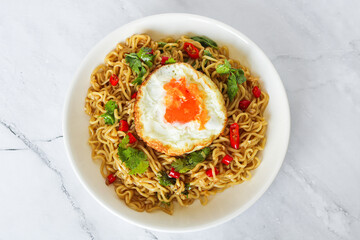 An overhead shot of a plate of spicy noodles, topped with a fried egg, fresh red chili, and coriander leaves.