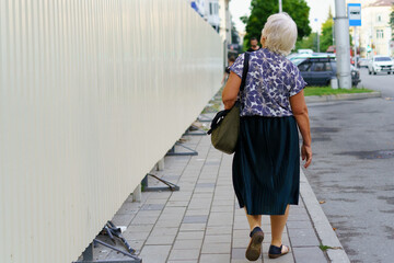 Elderly woman walking along a city sidewalk by a construction site on a sunny day