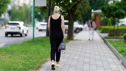 Woman walking on a city sidewalk during day near greenery, showcasing urban life and routine