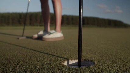 Female golfer places golf ball into hole on sunlit putting green - Powered by Adobe