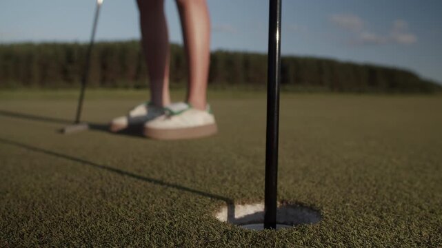 Female golfer places golf ball into hole on sunlit putting green