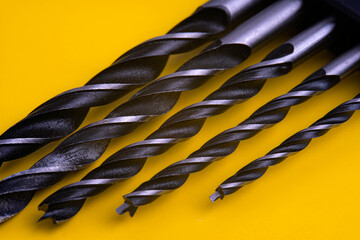 Assorted drill bits on a yellow background. Close-up showcases a set of various-sized metal drill bits arranged diagonally on a bright yellow surface. Highlighting their industrial design and utility.