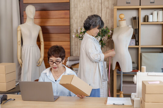 Adult asian daughter fashion designer working laptop while preparing ecommerce package box senior asian mother in background measuring mannequin inside boutique atelier teamwork small family business - Powered by Adobe