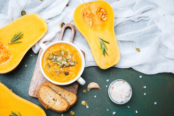 Bowl of squash soup with roasted seeds, fresh rosemary, and crusty bread slices on a dark surface.
