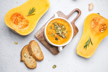 Butternut squash soup in bowl with seeds and herbs, next to squash halves and bread slices.