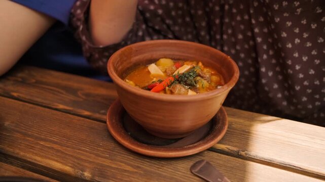 A person savors a bowl of lagman with beef featuring colorful vegetables while seated at a rustic wooden table in a charming restaurant during midday
