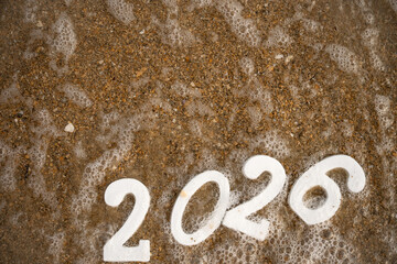 White numbers 2026, partially covered by sea foam, rest on a sandy beach, embodying the spirit of celebration for the upcoming new year while capturing a serene coastal scene