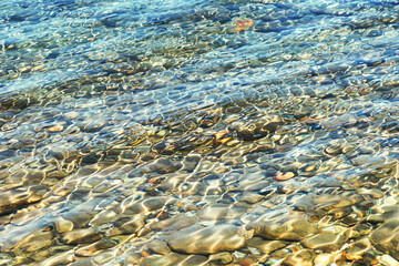 Clear shallow sea water over colorful stones with bright reflections and wave ripples in Halkidiki, Greece.