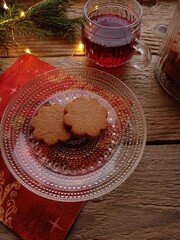 Close up of a few gingerbread cookies on a glass plate and in a jar. They are on a wooden table with a mug of mulled wine and some decorative elements.