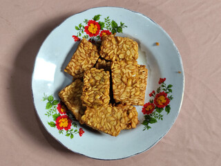 A pile of crispy fried tempeh that looks delicious and tasty, in a glass plate with a floral motif.